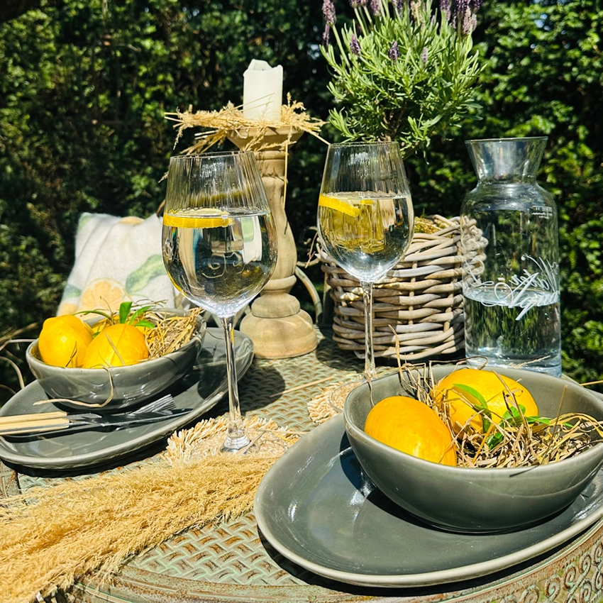 Gedeckter Gartentisch mit grauem Geschirr, Weingläsern mit Zitronenscheiben, Zitronen in Schalen, Glasflasche und Lavendelpflanze