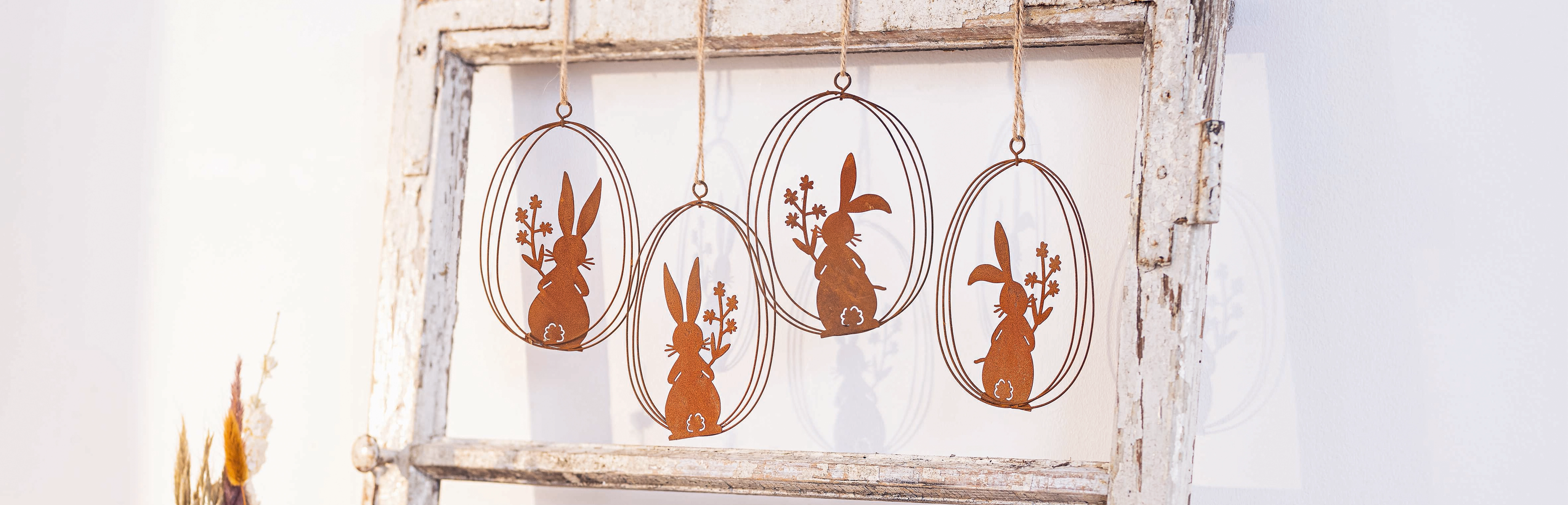 Osterdeko: rostfarbene Metall‑Hasenanhänger im ovalen Rahmen an weißer Holzleiter