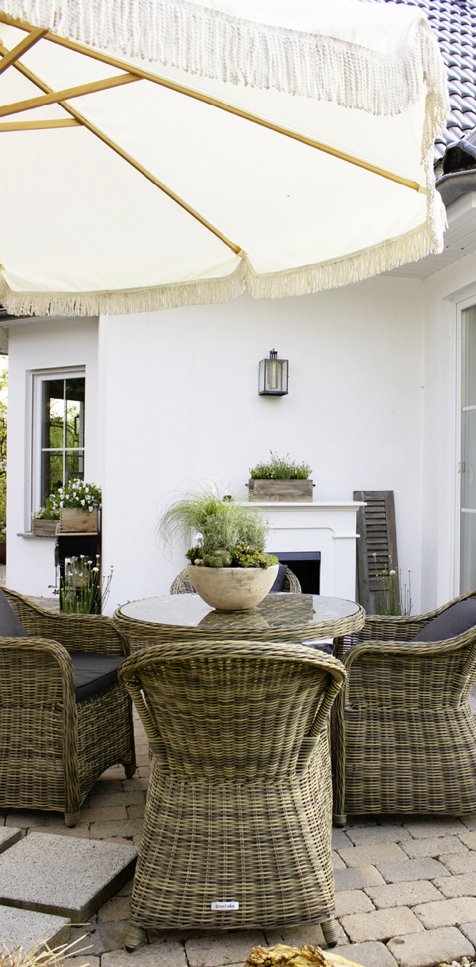 Terrasse mit runder Sitzgruppe aus Rattan, Sonnenschirm, bepflanzten Körben und Outdoor‑Kamin vor weißer Hausfassade