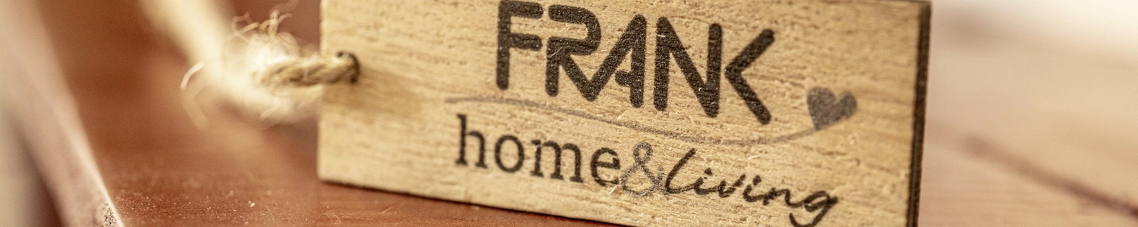 Holzschild mit eingraviertem Schriftzug ‚FRANK home & living‘ und Kordelaufhängung auf einer Holzoberfläche