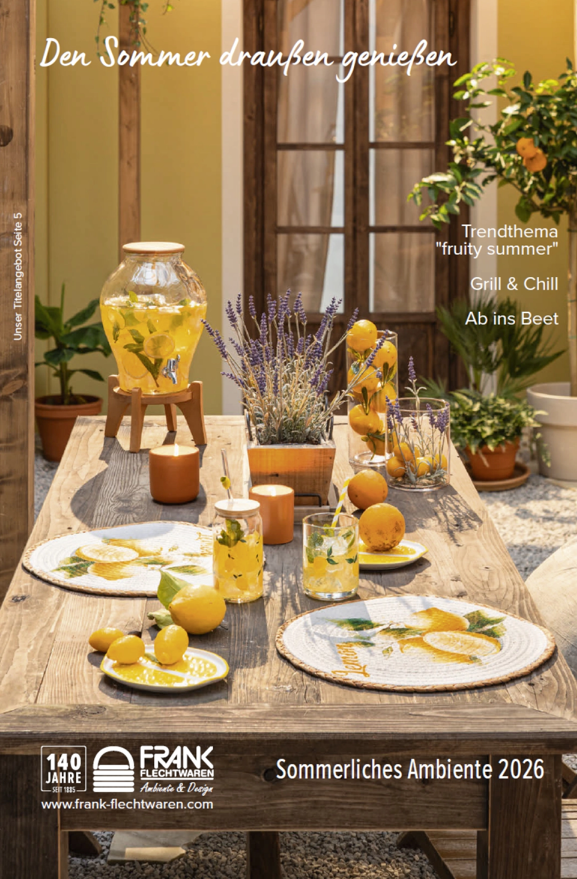 Sommerlich gedeckter Holztisch im Außenbereich mit Zitronen, Getränkespender, Gläsern, Kerzen, Lavendelpflanzen und Geschirr im Trendthema ‚fruity summer‘, Katalogmotiv Sommer 2026