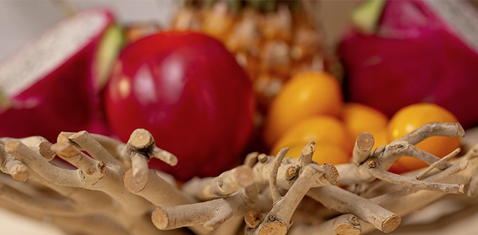 Tischdeko: Holzschale aus Zweigen mit roter Drachenfrucht, Apfel und gelben Früchten