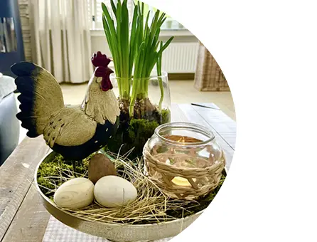 Osterdekoration auf Tablett mit Hahnfigur, Glaswindlicht, Eiern und Frühlingspflanzen im Wohnraum