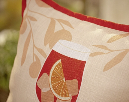 Dekokissen mit sommerlichem Motiv aus Orangenstücken in einem roten Glas auf hellem Stoff