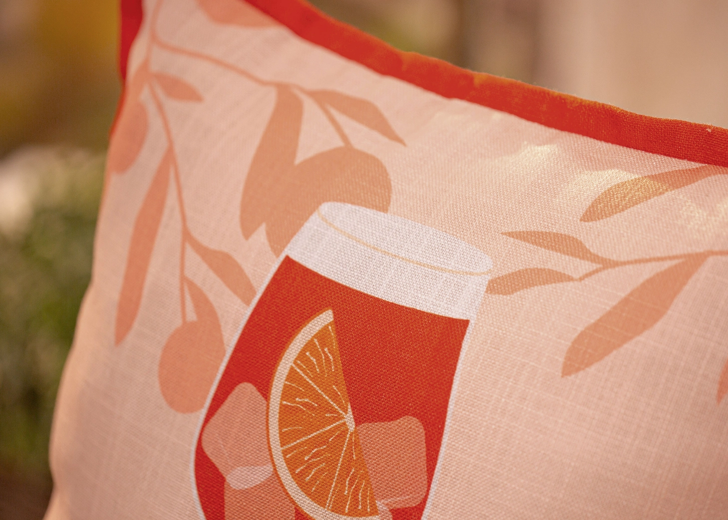 Detailaufnahme einer Kissenhülle mit sommerlichem Motiv aus rotem Getränkeglas, Orangenscheibe und floralem Muster in warmen Rosé‑ und Orangetönen