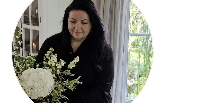 Person in Innenraum arrangiert einen weißen Blumenstrauß neben einem Fenster mit Blick ins Grüne