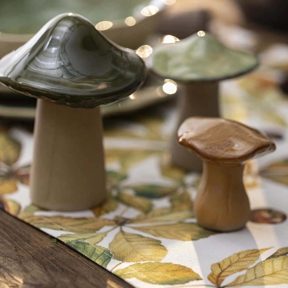 „Dekorative Pilzfiguren aus Holz und Glas auf einem Tischläufer mit herbstlichem Blättermuster als herbstliche Tischdekoration