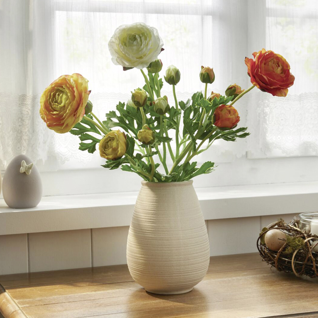 Ranunkelstrauß in warmen Farben in cremefarbener Vase am Fenster