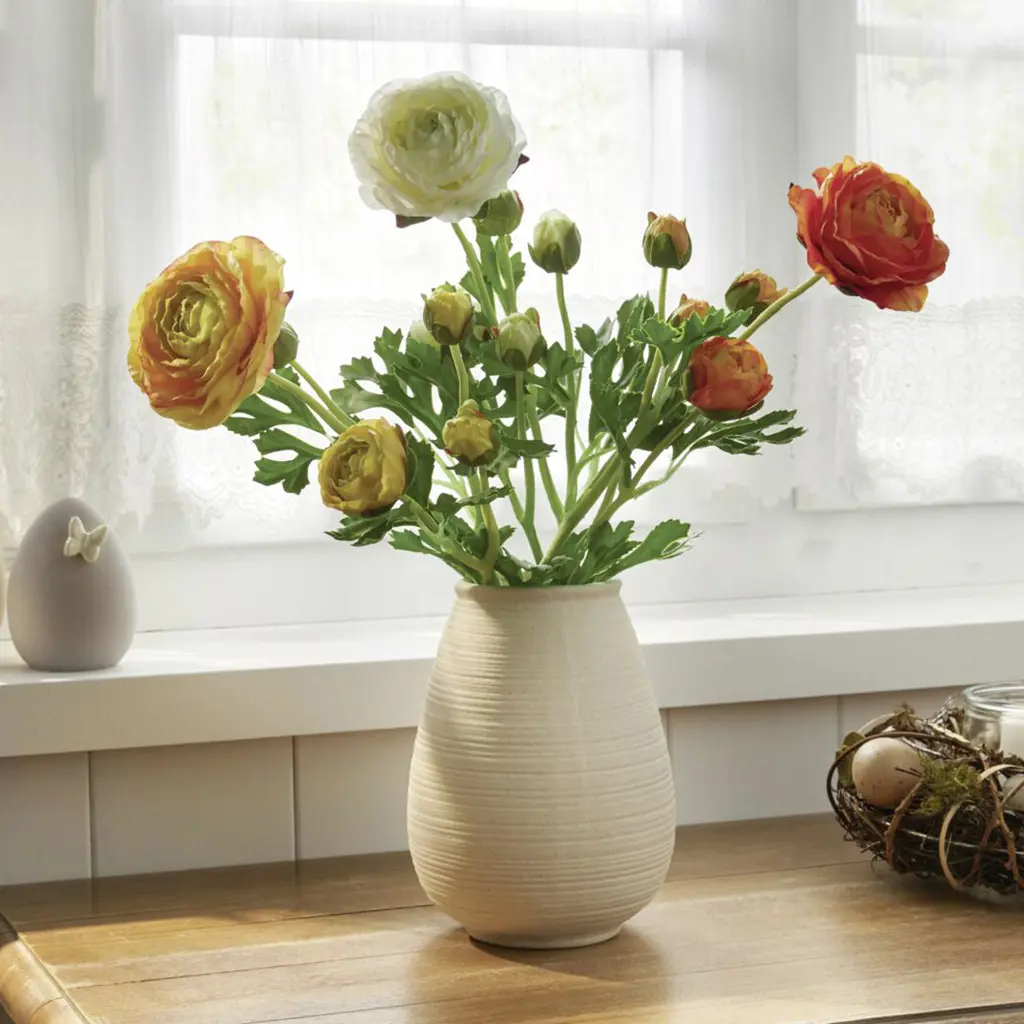 Ranunkelstrauß in warmen Farben in cremefarbener Vase am Fenster