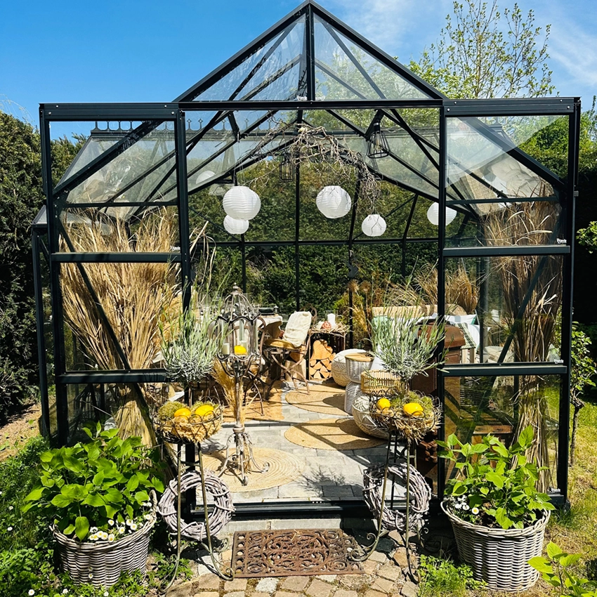 Dekoriertes Gewächshaus im Garten mit offenen Glastüren, bepflanzten Körben, Metallständern mit Zitronen, Laternen und Sitzbereich im Innenraum