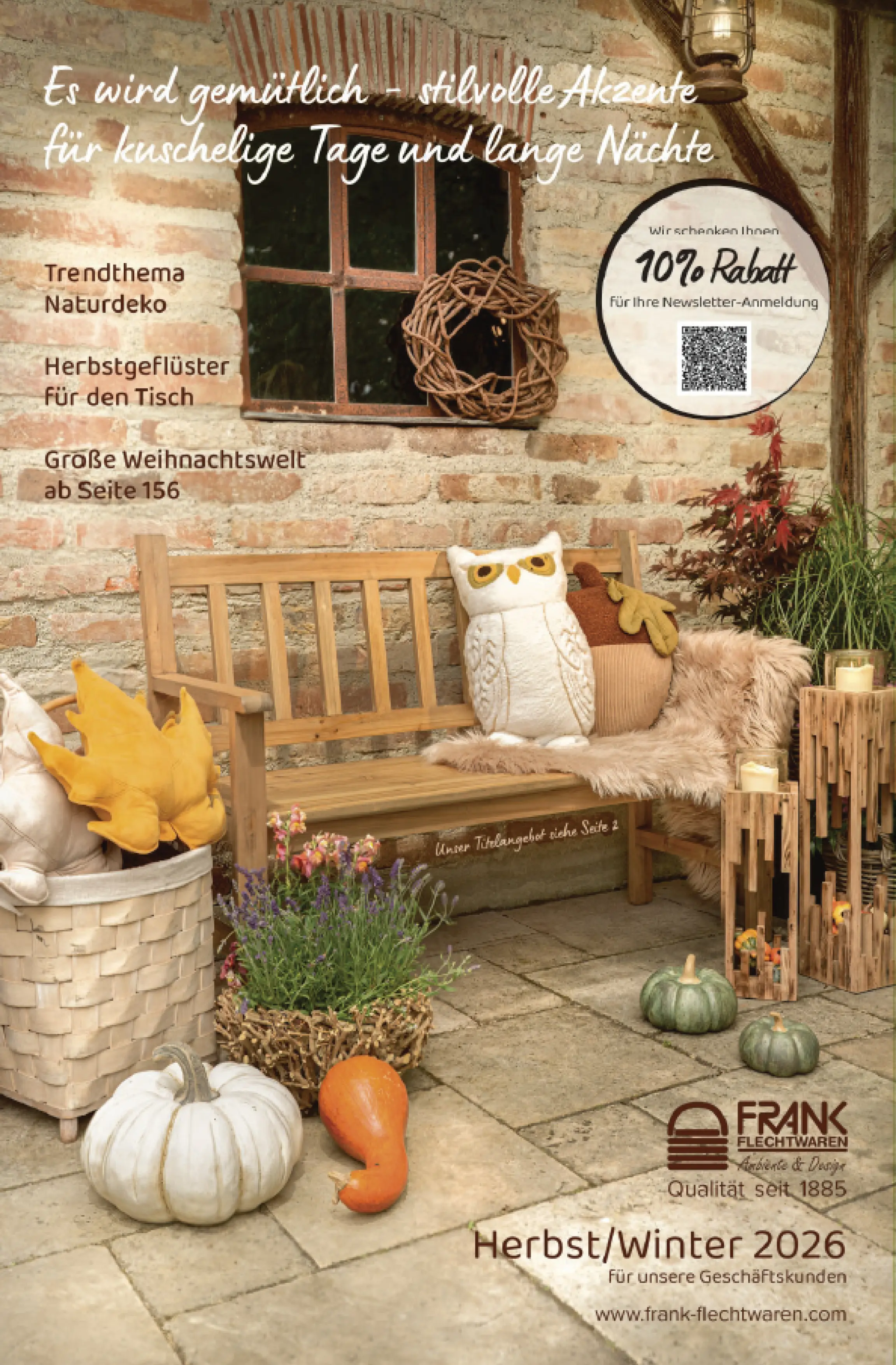 Herbstliche Outdoor-Dekoration mit Holzbank, Kürbissen, Eulenkissen, Holzwindlichtern, Flechtkorb und Pflanzen vor einer rustikalen Backsteinwand, Titelmotiv Katalog Herbst/Winter 2026 für Geschäftskunden