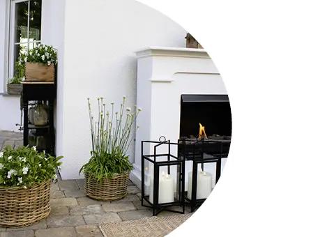 Terrassendekoration mit weißem Outdoor‑Kamin, schwarzen Metalllaternen mit Kerzen und bepflanzten Flechtkörben