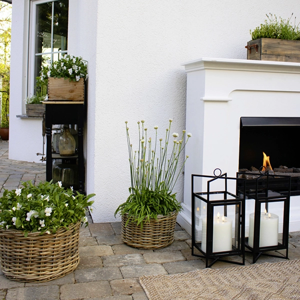 Terrassendekoration mit bepflanzten Flechtkörben, Holzpflanzkasten und schwarzen Laternen mit Kerzen vor Hauswand