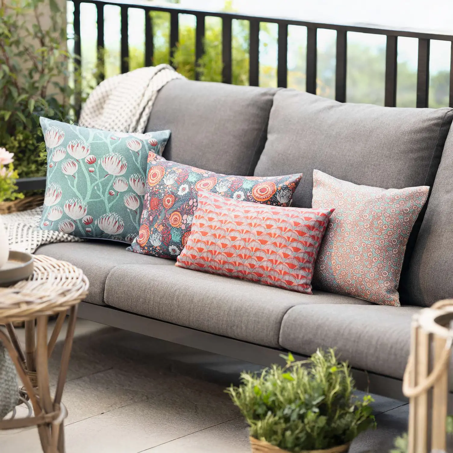 Graues Outdoor‑Sofa auf Terrasse mit dekorativen Kissen in Grün, Orange und bunten Mustern, dazu Pflanzen und Laternen