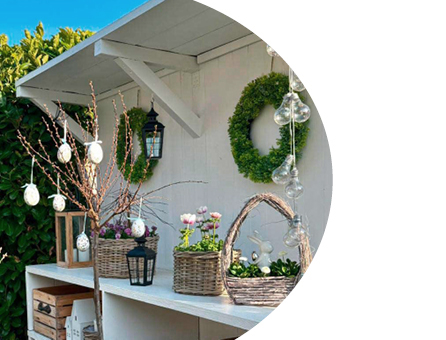 Frühlingshafte Osterdekoration auf weißem Regal mit bepflanzten Körben, Zweigen mit Ostereiern, Kranz und Laternen im Gartenbereich