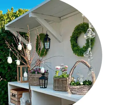 Frühlingshafte Osterdekoration auf weißem Regal mit bepflanzten Körben, Zweigen mit Ostereiern, Kranz und Laternen im Gartenbereich